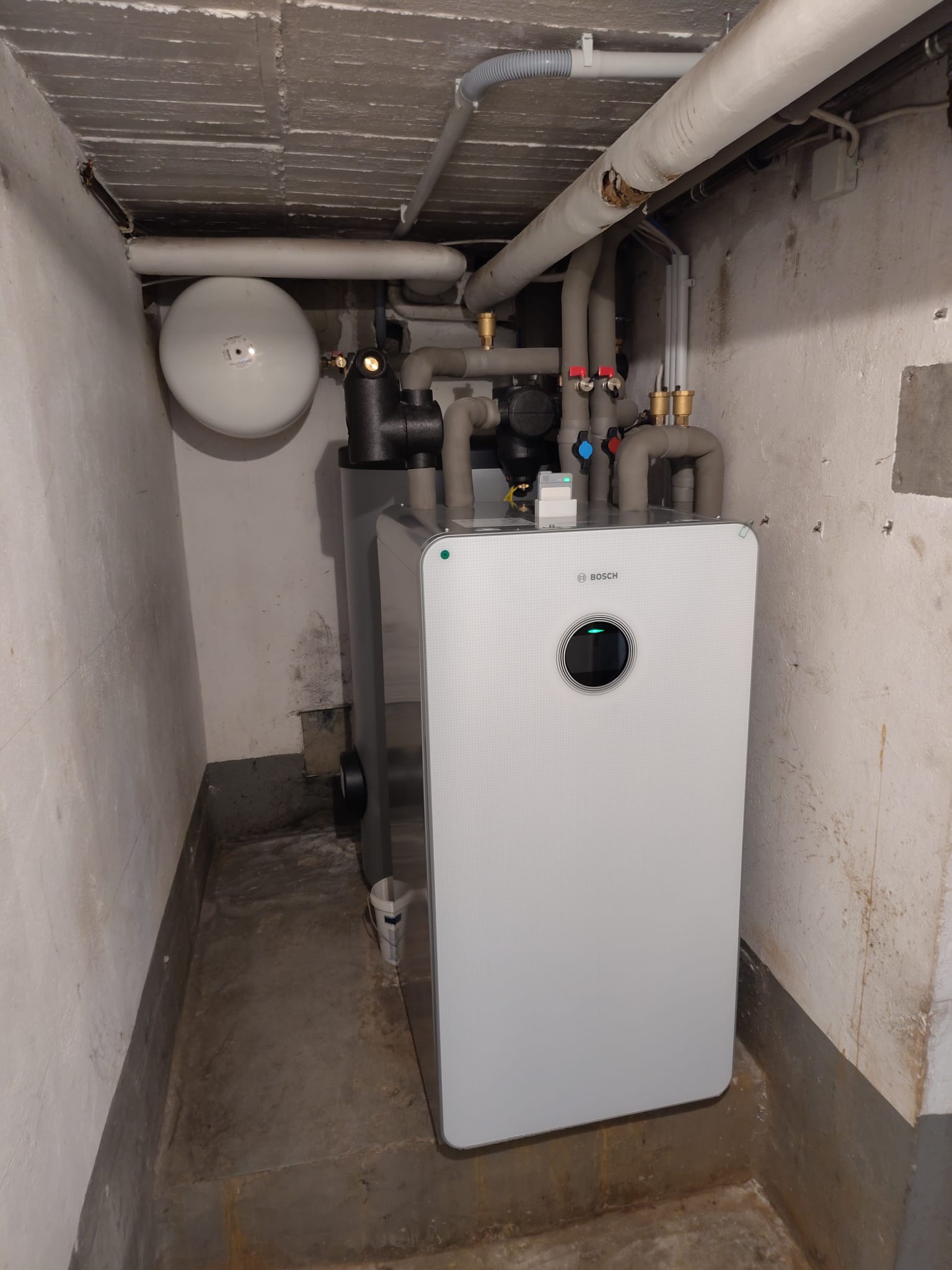 Bosch-Heizungseinheit in engem Kellerraum mit vielen isolierten Rohrleitungen und Wandarmaturen.